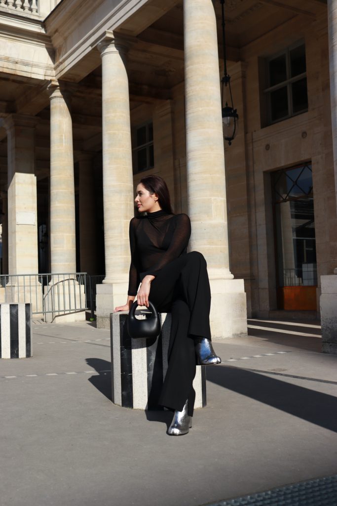 Fashion blogger and stylist Alina Barchet poses for a photo shot at Colonnes de Buren/Les Deux Plateaux in Paris. Alina Barchet styled a black slightly see-through long sleeve turtle neck top with black pants, a black leather bag and silver tabi boots from Maison Margiela.